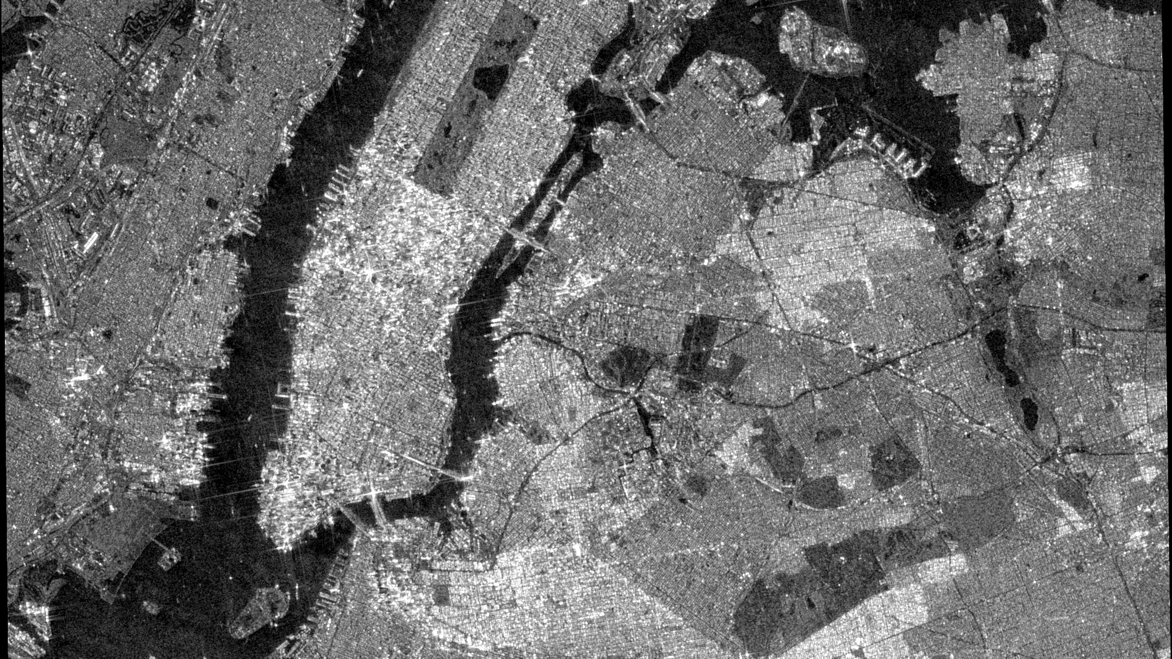 Black and white radar satellite image of Manhattan, New York, with the island glowing bright between dark surrounding rivers.