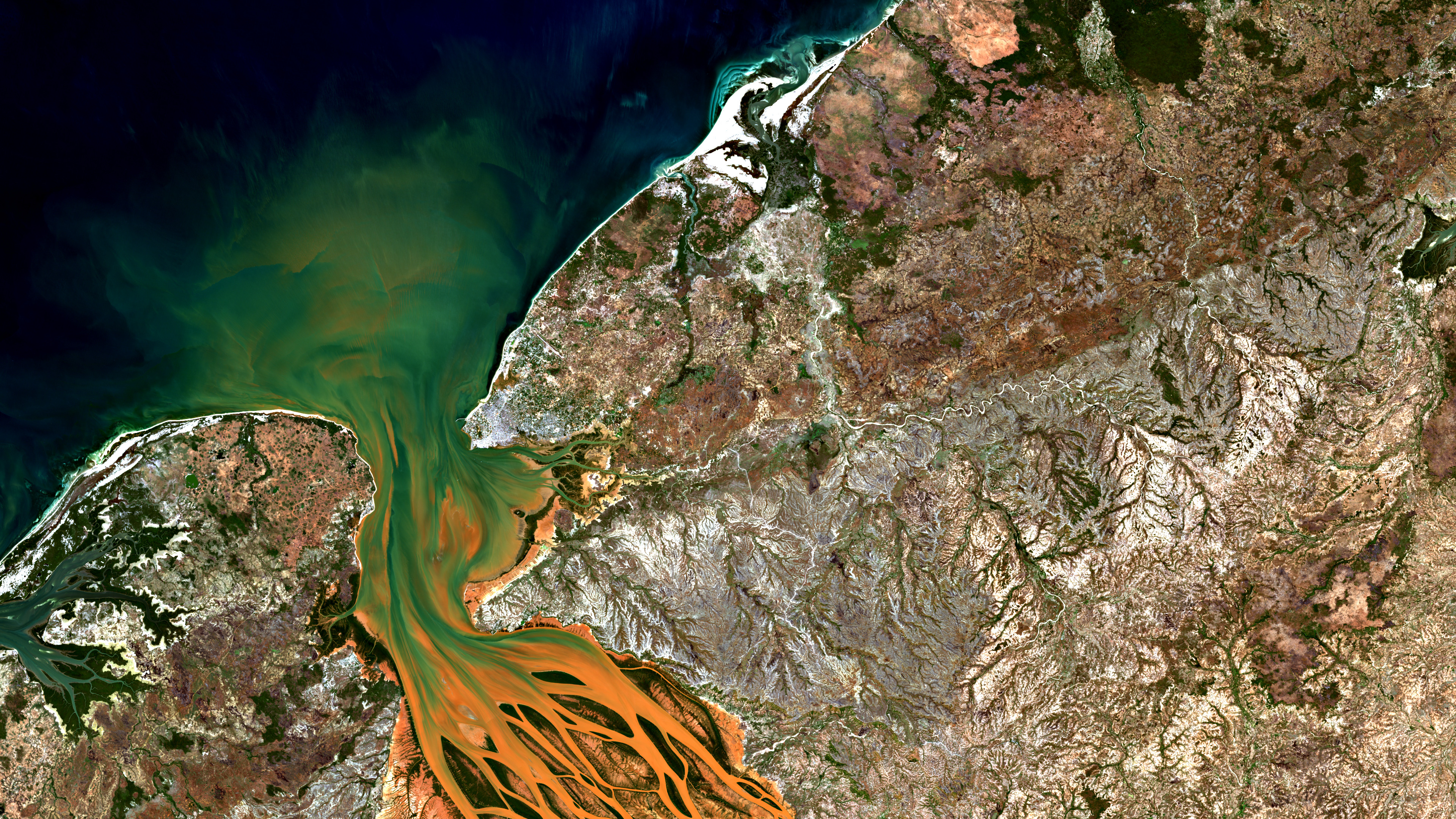 A wide satellite view of Madagascar’s Betsiboka estuary with orange braided sediment channels entering green coastal water beside dark ocean and rugged brown highlands.