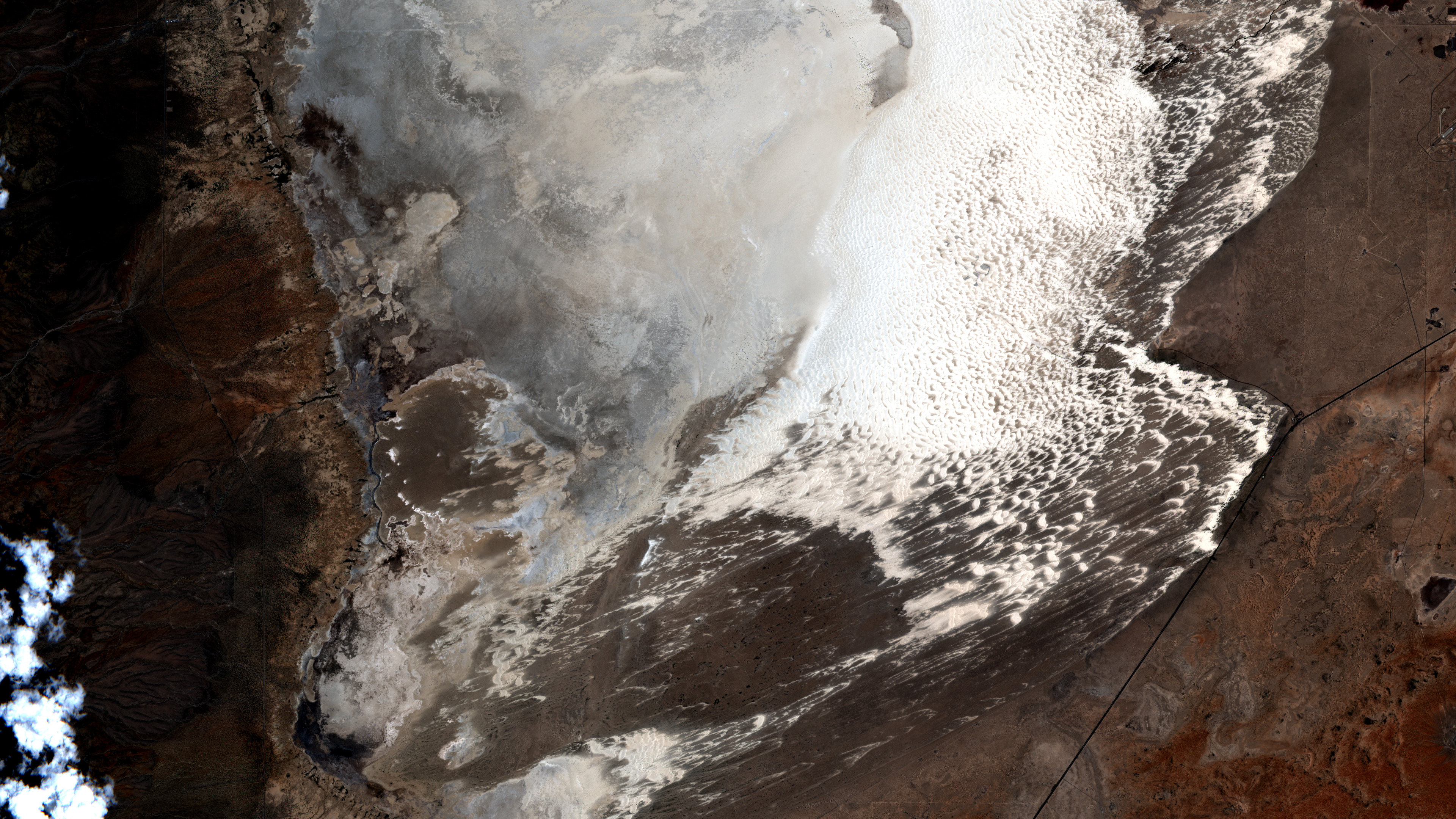 Satellite view of a large white gypsum dune field surrounded by brown desert, dark mountain shadows, roads, and a few small clouds.