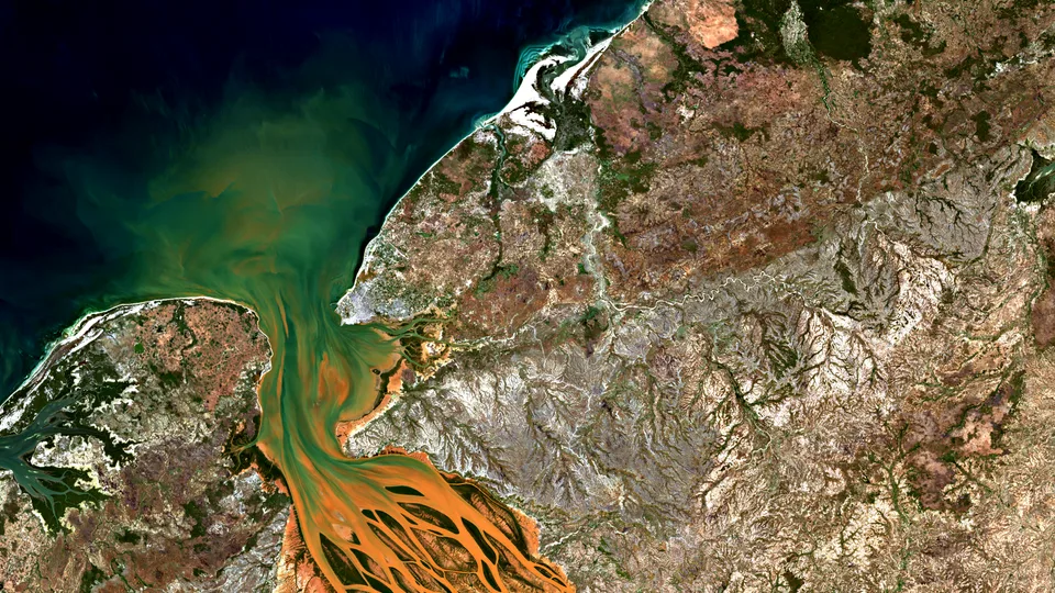 A wide satellite view of Madagascar’s Betsiboka estuary with orange braided sediment channels entering green coastal water beside dark ocean and rugged brown highlands.