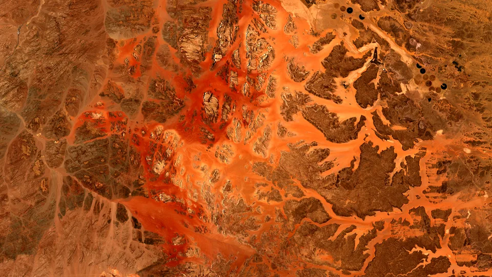 Satellite image of Wadi Rum showing reddish sandstone massifs, pale branching desert wadis, darker rock plateaus, roads, and small green agricultural plots.